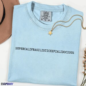 Puede incluir: Una camiseta azul claro con la palabra "SUPERCALIFRAGILISTICEXPIALIDOCIOUS" impresa en negro. Un collar con colgante de estrella dorada descansa sobre la camiseta. La camiseta tiene una etiqueta Comfort Colors, talla XL-TG-EG.