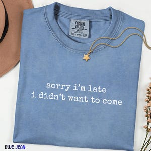 May include: A blue short-sleeved t-shirt with the white text "sorry i'm late i didn't want to come" printed on the front. The shirt features a Comfort Colors brand label at the inner neckline, indicating a size XL, and is made of 100% cotton.