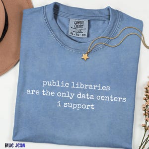May include: A blue jean colored t-shirt with the white text "public libraries are the only data centers i support" printed on the front. The shirt features a Comfort Colors brand label at the neckline and is paired with a gold star pendant necklace.