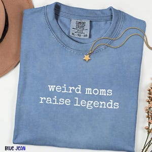 May include: A blue jean colored t-shirt with the text "weird moms raise legends" in white. A gold star pendant necklace rests on the shirt. The shirt has a Comfort Colors tag.
