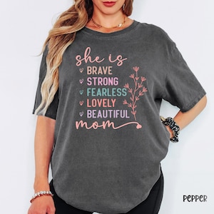 May include: Dark gray t-shirt with the words "she is" in pink script, followed by "brave, strong, fearless, lovely, beautiful mom" in pink and teal. A floral design is on the right side of the text.