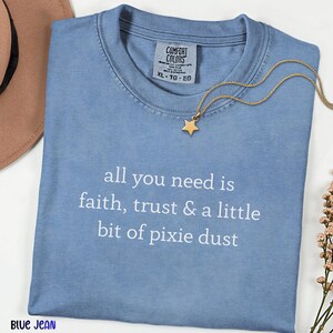 May include: A blue jean colored t-shirt with the white text all you need is faith, trust & a little bit of pixie dust printed on the front. The shirt features a Comfort Colors brand label at the neckline and a gold star pendant necklace.