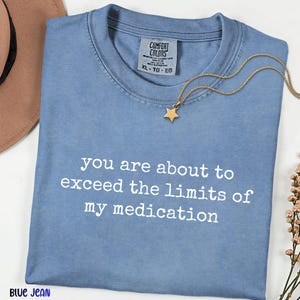 May include: A blue crew neck t-shirt with the white text you are about to exceed the limits of my medication printed on the front. The shirt features a Comfort Colors label at the neckline and is paired with a gold star pendant necklace.