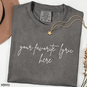 May include: A gray Comfort Colors t-shirt with the text "your favorite lyric here" in white script. A gold star pendant necklace rests on the shirt. The shirt is size XL-TG-EG.