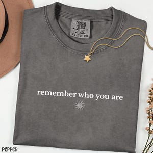May include: A charcoal gray crew neck t-shirt featuring the white text "remember who you are" above a small sun icon. The shirt has a Comfort Colors label at the inner neckline and is paired with a gold-toned necklace with a star pendant.