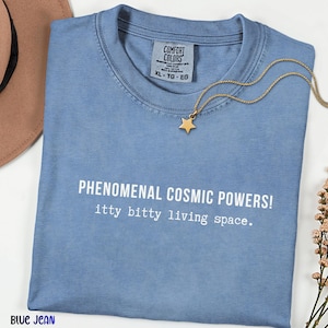 May include: A blue jean colored t-shirt with a crew neckline and a Comfort Colors label. The shirt features the white printed text: PHENOMENAL COSMIC POWERS! itty bitty living space. A gold-toned star pendant necklace rests on the fabric near the collar.