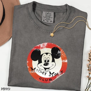 May include: A gray Comfort Colors t-shirt featuring a distressed Mickey Mouse Club graphic in a red and white circle. A gold star pendant necklace rests on the shirt. The shirt is size XL-TG-EG.