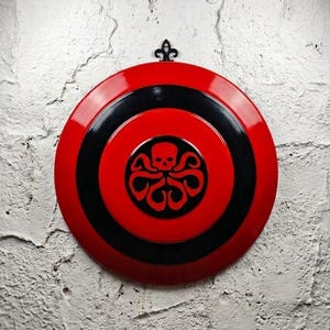 Puede incluir: Un escudo redondo rojo y negro con un dise&ntilde;o de calavera y pulpo en el centro. El escudo est&aacute; montado en una pared de ladrillos blancos. Una pieza decorativa de metal negro est&aacute; en la parte superior.