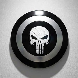 K&ouml;nnte beinhalten: Ein schwarzer und silberner Schild mit einem wei&szlig;en Totenkopf-Emblem. Der Schild hat ein kreisf&ouml;rmiges Design mit einem schwarzen Au&szlig;enring, einem silbernen Mittelring und einem schwarzen Zentrum mit dem Totenkopf. Der Totenkopf hat ein rissiges Aussehen.