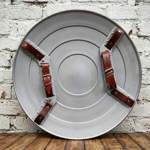 May include: A gray, circular shield with brown leather straps and silver metal accents. The shield has concentric circles and a red border. The straps are secured with buckles. The shield is displayed against a white brick wall.