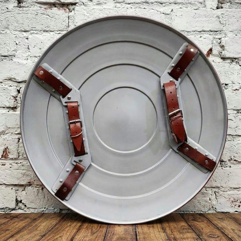 May include: A gray, circular shield with brown leather straps and silver metal accents. The shield has concentric circles and a red border. The straps are secured with buckles. The shield is displayed against a white brick wall.