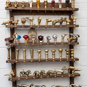 Könnte beinhalten: Ein hölzernes Displayregal mit einer Sammlung dekorativer Stockgriffe. Die Griffe weisen verschiedene Designs auf, darunter Schädel, Tierfiguren und abstrakte Formen. Das Regal ist aus dunklem Holz gefertigt und an einer weißen Backsteinmauer befestigt. Jeder Griff ist nummeriert.