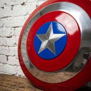 May include: A red, white, and blue shield with a silver star in the center. The shield has a glossy finish and a circular design. The shield is resting on a wooden surface.