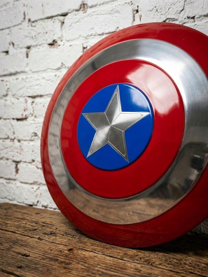 May include: A red, white, and blue shield with a silver star in the center. The shield has a glossy finish and a circular design. The shield is resting on a wooden surface.