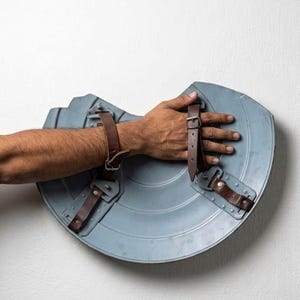 May include: A light blue, semi-circular shield with brown leather straps. The shield has a metallic appearance and is designed with a series of concentric circles. The straps are riveted to the shield, suggesting a durable construction.