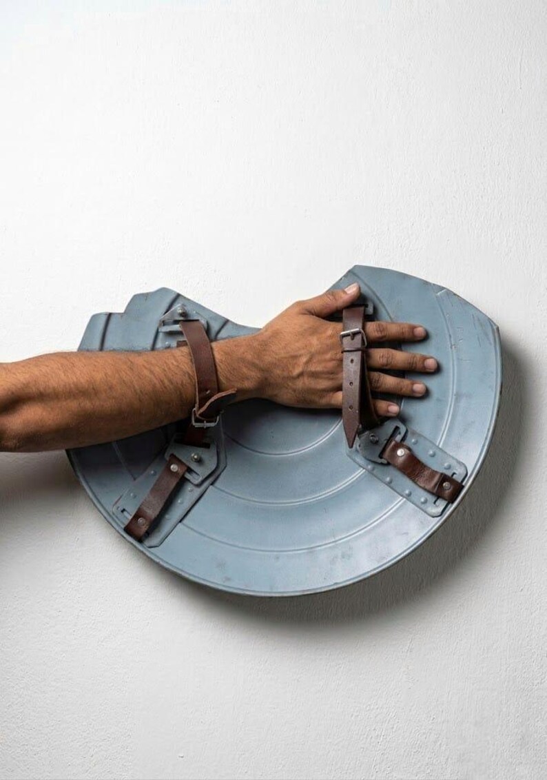 May include: A light blue, semi-circular shield with brown leather straps. The shield has a metallic appearance and is designed with a series of concentric circles. The straps are riveted to the shield, suggesting a durable construction.