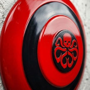 Puede incluir: Un escudo rojo y negro brillante con un emblema central que presenta una calavera y tent&aacute;culos. El escudo tiene c&iacute;rculos conc&eacute;ntricos rojos y negros, con el emblema en el centro. El dise&ntilde;o es audaz e impactante.