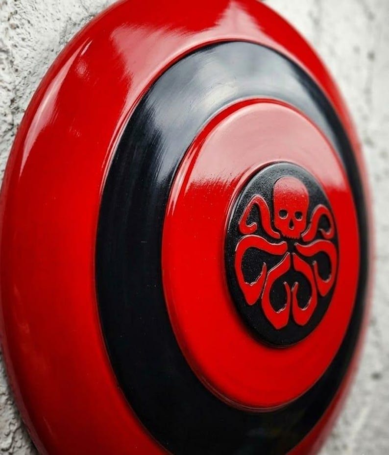 Puede incluir: Un escudo rojo y negro brillante con un emblema central que presenta una calavera y tent&aacute;culos. El escudo tiene c&iacute;rculos conc&eacute;ntricos rojos y negros, con el emblema en el centro. El dise&ntilde;o es audaz e impactante.