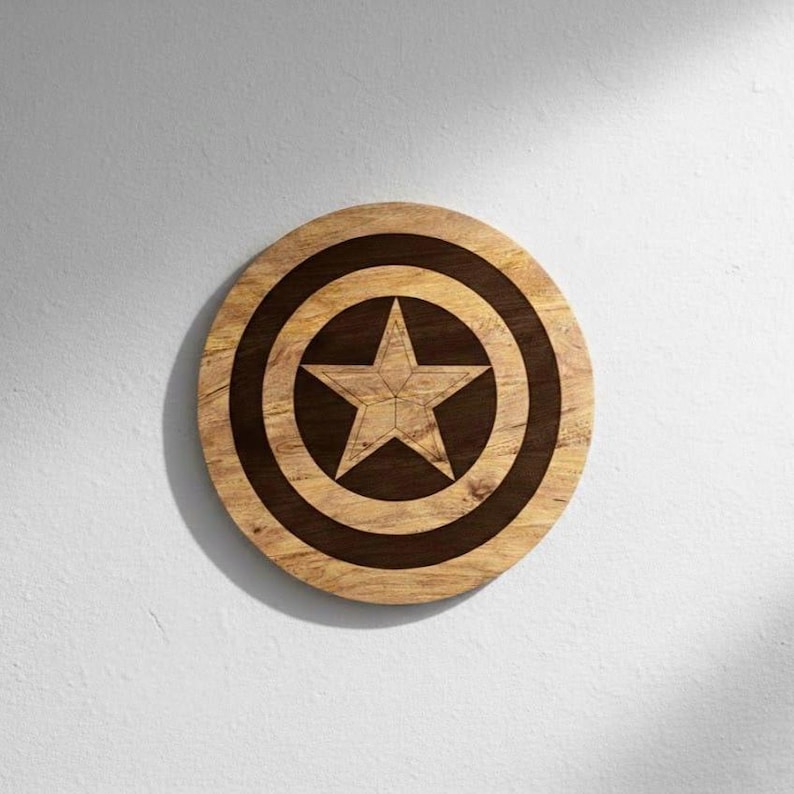 May include: A round wooden shield with a star design. The shield is made of light brown wood with a dark brown star in the center and a dark brown ring around the star. The shield is a decorative item.