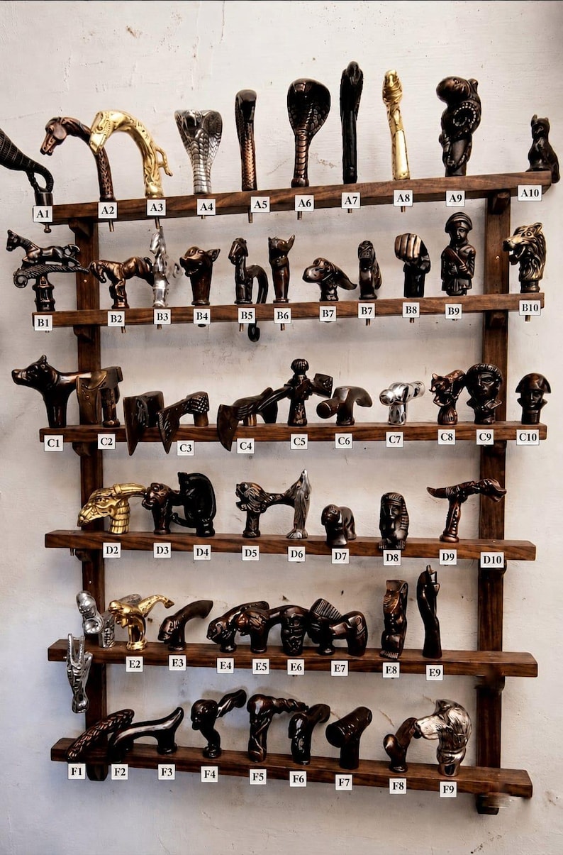 May include: A wooden wall-mounted display rack featuring six horizontal shelves holding numerous decorative cane handles in various metallic and dark finishes, each labelled with alphanumeric codes from A1 to F9 for identification.