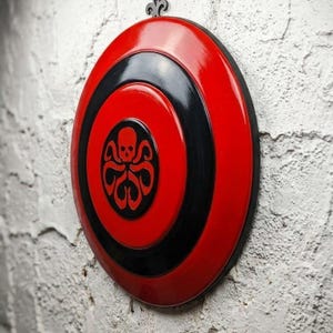 Puede incluir: Un escudo rojo y negro con acabado brillante, con un emblema central de una calavera y tent&aacute;culos. El escudo est&aacute; montado en una pared blanca texturizada, mostrando un dise&ntilde;o decorativo.