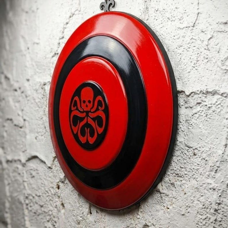 Puede incluir: Un escudo rojo y negro con acabado brillante, con un emblema central de una calavera y tent&aacute;culos. El escudo est&aacute; montado en una pared blanca texturizada, mostrando un dise&ntilde;o decorativo.