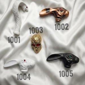 May include: Five decorative cane handles labelled with numbers. 1001 is a white snake, 1002 is a copper-toned rabbit with red eyes, 1003 is a gold skull with red eyes, 1004 is a white rabbit with red eyes, and 1005 is a black rabbit with green eyes.