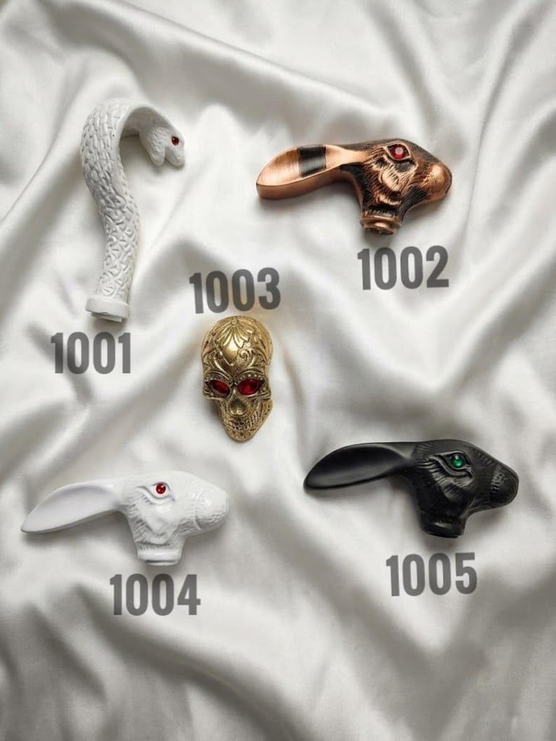 May include: Five decorative cane handles labelled with numbers. 1001 is a white snake, 1002 is a copper-toned rabbit with red eyes, 1003 is a gold skull with red eyes, 1004 is a white rabbit with red eyes, and 1005 is a black rabbit with green eyes.