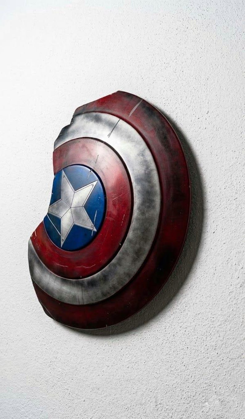 May include: A distressed Captain America shield, featuring a blue central disc with a white star, surrounded by concentric rings of red, silver, and dark red. The shield has a battle-worn appearance, with visible damage.