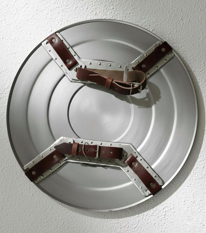 Puede incluir: Un escudo redondo de color plateado con correas de cuero marr&oacute;n y detalles met&aacute;licos. El escudo tiene un acabado de metal cepillado y un dise&ntilde;o circular central. Las correas est&aacute;n remachadas al escudo y se ve una hebilla.