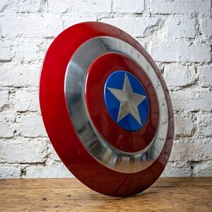 May include: A red, white, and blue shield with a silver star in the center. The shield is round with a red outer ring, a silver middle ring, and a blue center. The shield is made of metal and is likely a collectible or a prop.