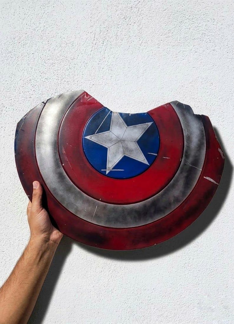 May include: A distressed Captain America shield, featuring a red, white, and blue color scheme. The shield has a central blue circle with a white star, surrounded by red and silver concentric rings. The shield has a damaged appearance.
