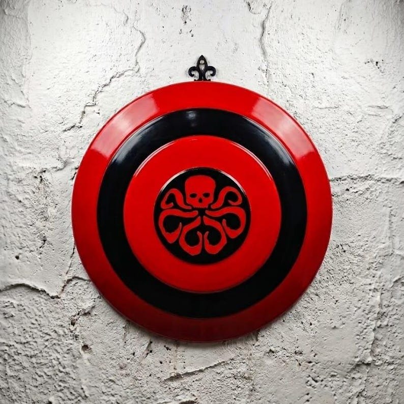 Puede incluir: Un escudo redondo rojo y negro con un dise&ntilde;o de calavera y pulpo en el centro. El escudo est&aacute; montado en una pared de ladrillos blancos. Una pieza decorativa de metal negro est&aacute; en la parte superior.