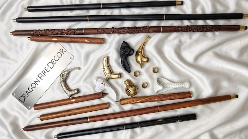 May include: A collection of walking stick components including long black and brown wooden shafts, various ornate metal and white handles, small brass rings, and a rectangular metal plate with the text DRAGON FIRE DECOR engraved on it.