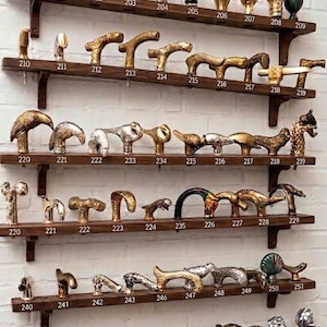 May include: A collection of various metal cane handles displayed on six wooden shelves. Each handle is numbered from 200 to 265, featuring diverse shapes including animal heads, abstract forms, and spherical knobs in gold, silver, and bronze tones.