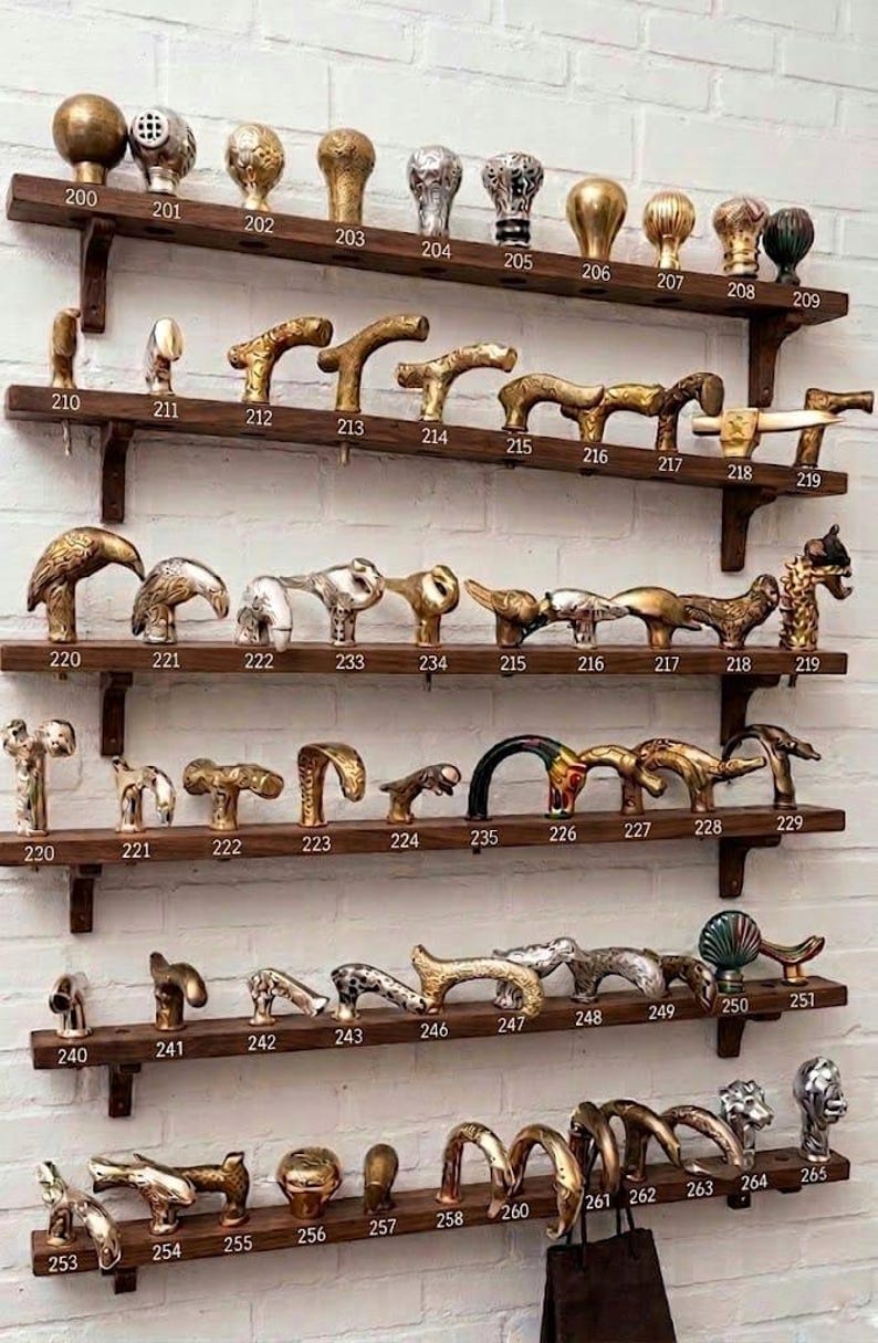 May include: A collection of various metal cane handles displayed on six wooden shelves. Each handle is numbered from 200 to 265, featuring diverse shapes including animal heads, abstract forms, and spherical knobs in gold, silver, and bronze tones.
