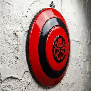 Puede incluir: Decoraci&oacute;n de pared circular roja y negra con un emblema de calavera y pulpo. El dise&ntilde;o presenta c&iacute;rculos conc&eacute;ntricos rojos y negros, con un borde negro. Una pieza met&aacute;lica decorativa est&aacute; en la parte superior para colgar.