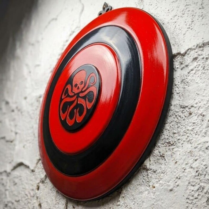 Puede incluir: Un escudo rojo y negro con un dise&ntilde;o en espiral. El escudo presenta un emblema central con una calavera y tent&aacute;culos. El escudo est&aacute; montado en una pared blanca texturizada.