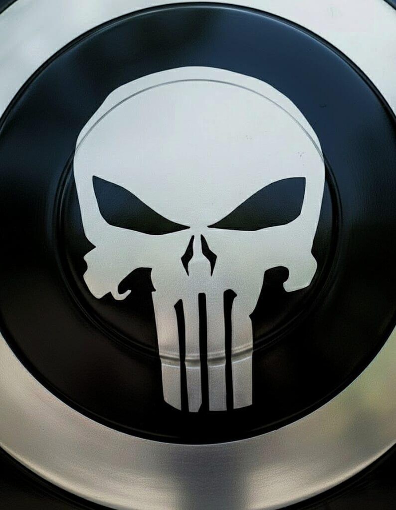 K&ouml;nnte beinhalten: Ein schwarz-silberner Schild mit einem wei&szlig;en Totenkopf-Emblem. Der Totenkopf hat scharfe, kantige Merkmale und ist auf dem Schild zentriert. Der Schild hat einen silbernen Rand und ein schwarzes Innendesign.