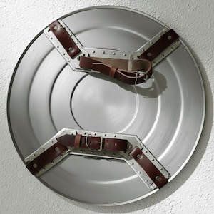 K&ouml;nnte beinhalten: Ein silberfarbener, runder Schild mit braunen Lederriemen und Metallakzenten. Der Schild hat ein zentrales kreisf&ouml;rmiges Design und ist wahrscheinlich ein Kost&uuml;mzubeh&ouml;r oder ein dekorativer Gegenstand.