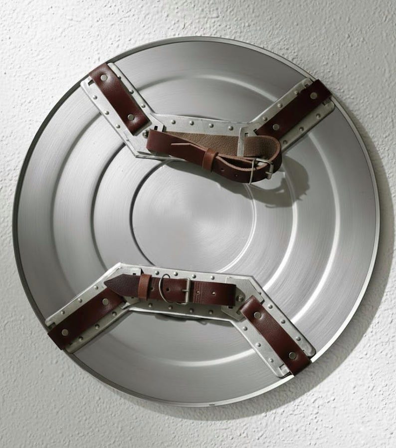 K&ouml;nnte beinhalten: Ein silberfarbener, runder Schild mit braunen Lederriemen und Metallakzenten. Der Schild hat ein zentrales kreisf&ouml;rmiges Design und ist wahrscheinlich ein Kost&uuml;mzubeh&ouml;r oder ein dekorativer Gegenstand.