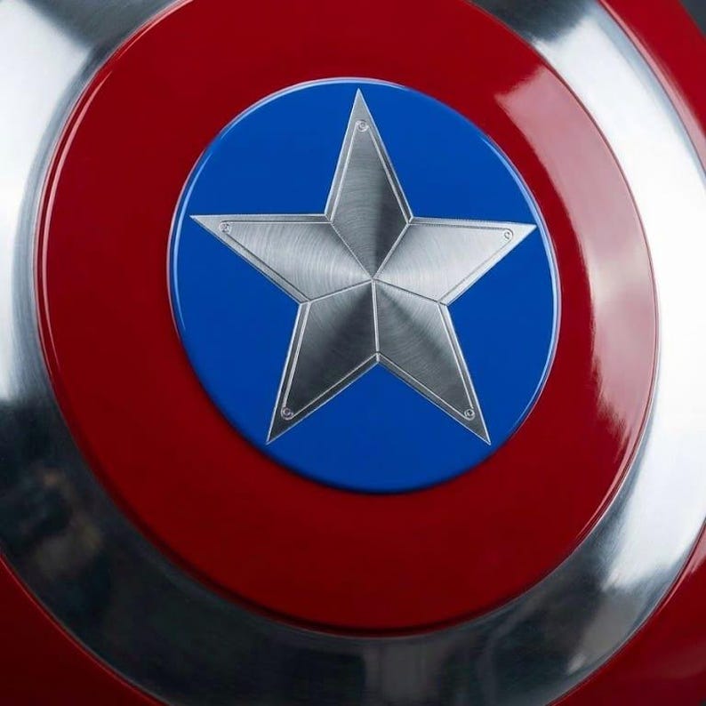 May include: A close-up of a shield with a red outer ring, a silver inner ring, and a blue center. A large, silver, five-pointed star is in the center of the blue circle. The shield has a metallic sheen.