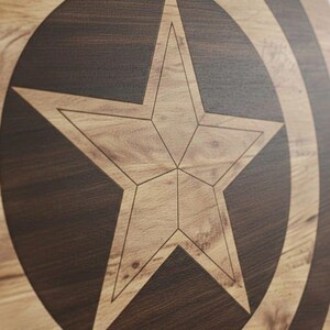 May include: Wooden shield featuring a large, five-pointed star in the center, with a dark brown circular border and concentric arcs. The star and arcs are lighter in color, creating a contrast against the darker wood grain.