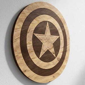 May include: A wooden shield with a star design. The shield is round with concentric circles in light and dark brown. A five-pointed star is in the center. The shield is mounted on a white wall.