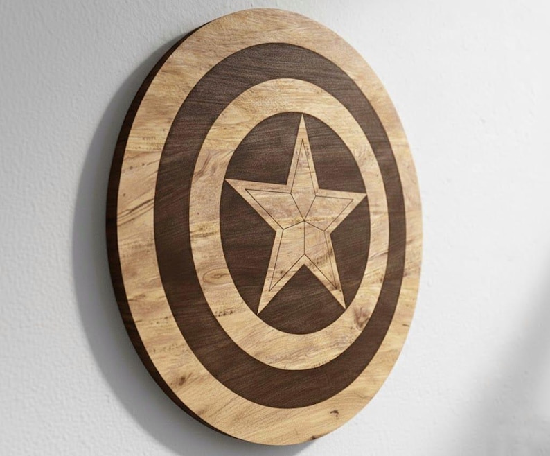 May include: A wooden shield with a star design. The shield is round with concentric circles in light and dark brown. A five-pointed star is in the center. The shield is mounted on a white wall.