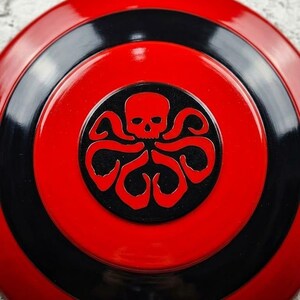 Puede incluir: Un escudo redondo rojo y negro con un emblema central. El emblema es una calavera negra con tent&aacute;culos, sobre un c&iacute;rculo negro. El escudo tiene anillos conc&eacute;ntricos rojos y negros.