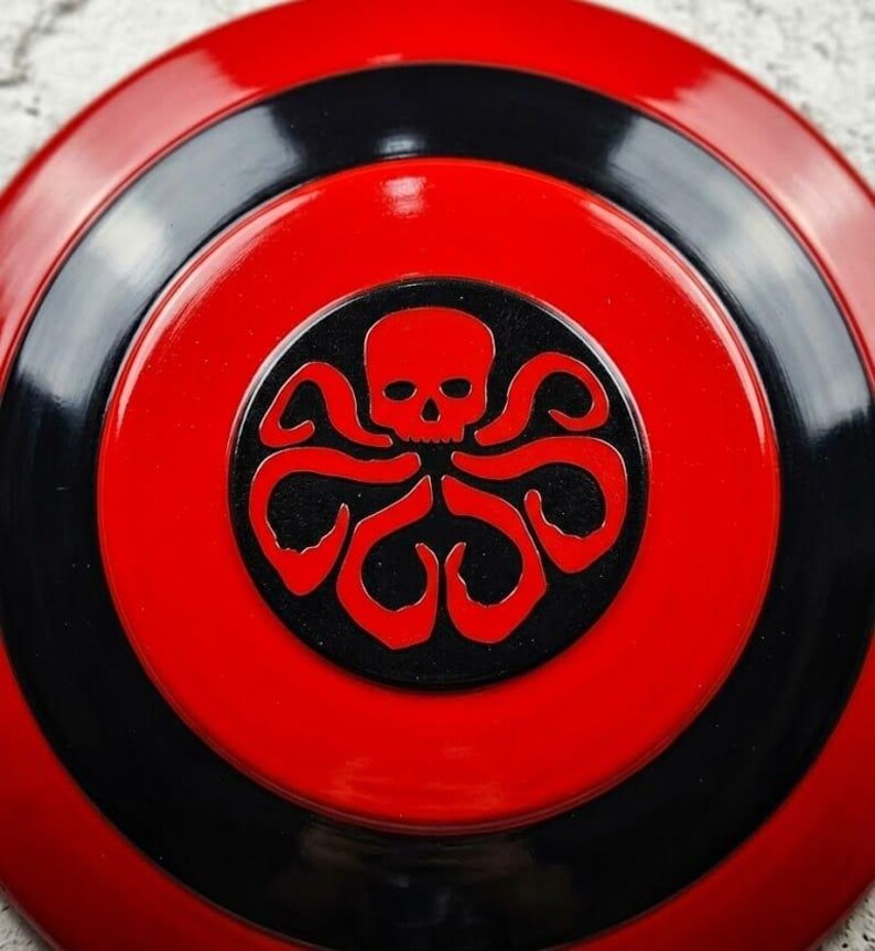 Puede incluir: Un escudo redondo rojo y negro con un emblema central. El emblema es una calavera negra con tent&aacute;culos, sobre un c&iacute;rculo negro. El escudo tiene anillos conc&eacute;ntricos rojos y negros.