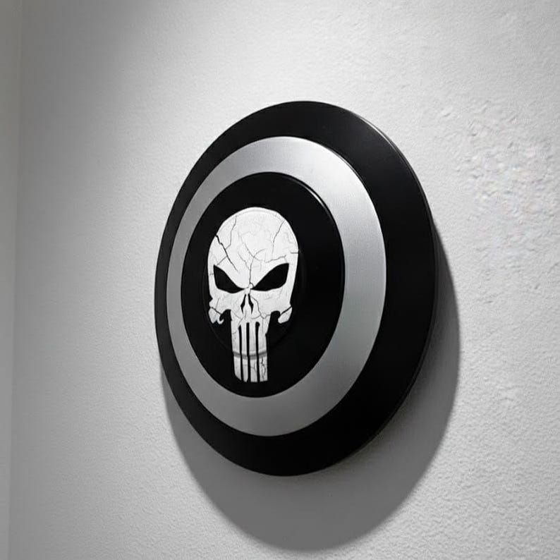 K&ouml;nnte beinhalten: Ein schwarz-silberner Schild mit einem wei&szlig;en Totenkopf-Emblem. Der Schild hat ein kreisf&ouml;rmiges Design mit einem schwarzen Au&szlig;enring, einem silbernen Mittelring und einem schwarzen Zentrum mit dem Totenkopf. Der Totenkopf hat ein rissiges Aussehen.