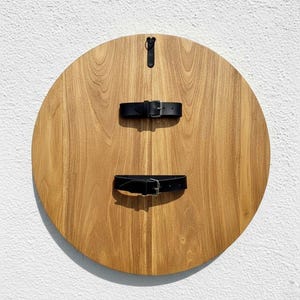 May include: A round wooden shield with a natural wood grain finish. Two black leather straps with silver buckles are attached to the front. A black leather strap is attached to the back for hanging.