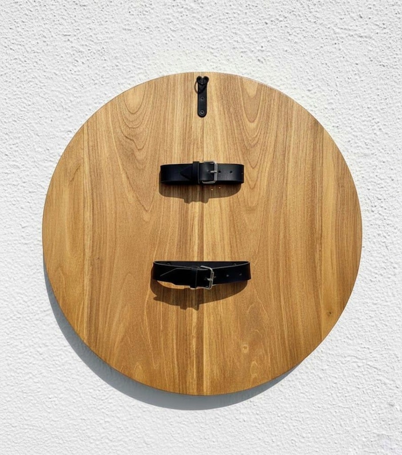 May include: A round wooden shield with a natural wood grain finish. Two black leather straps with silver buckles are attached to the front. A black leather strap is attached to the back for hanging.